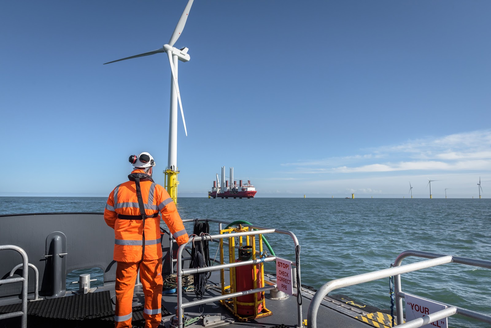 A person in orange safety gear stands on a boat looking at an offshore wind turbine and a distant platform at sea under a clear blue sky.