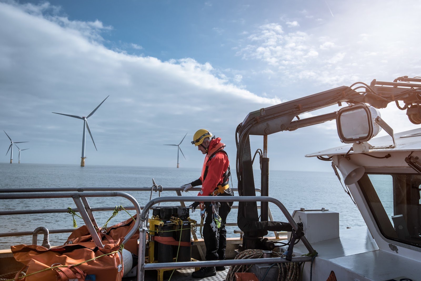 A worker in safety gear stands on a boat with equipment, near offshore wind turbines under a partly cloudy sky. The sea is calm, and the sun is shining in the background.