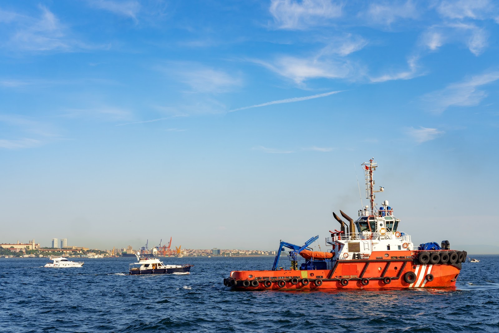A red and white tugboat sails on the water near several smaller boats, with a city skyline and cranes visible in the distance under a blue sky with wispy clouds.