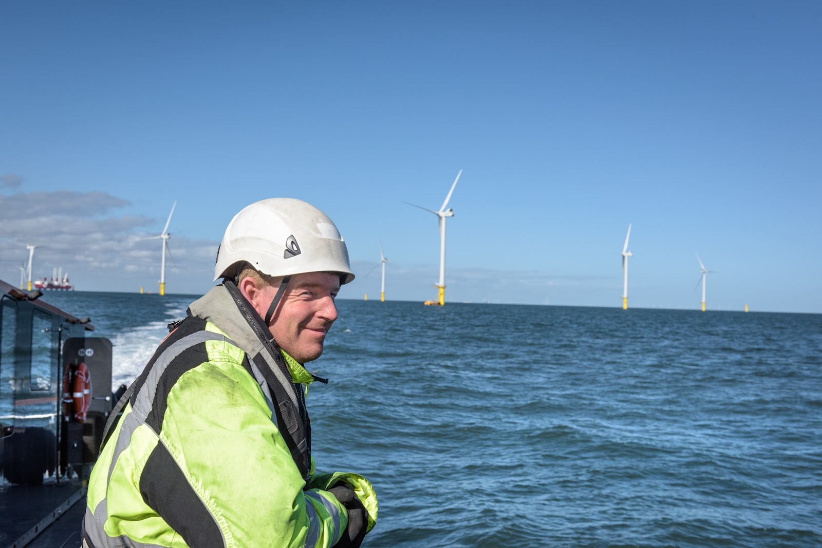 A person in a high-visibility jacket and hard hat stands on a boat, smiling, with offshore wind turbines visible in the ocean under a clear blue sky.