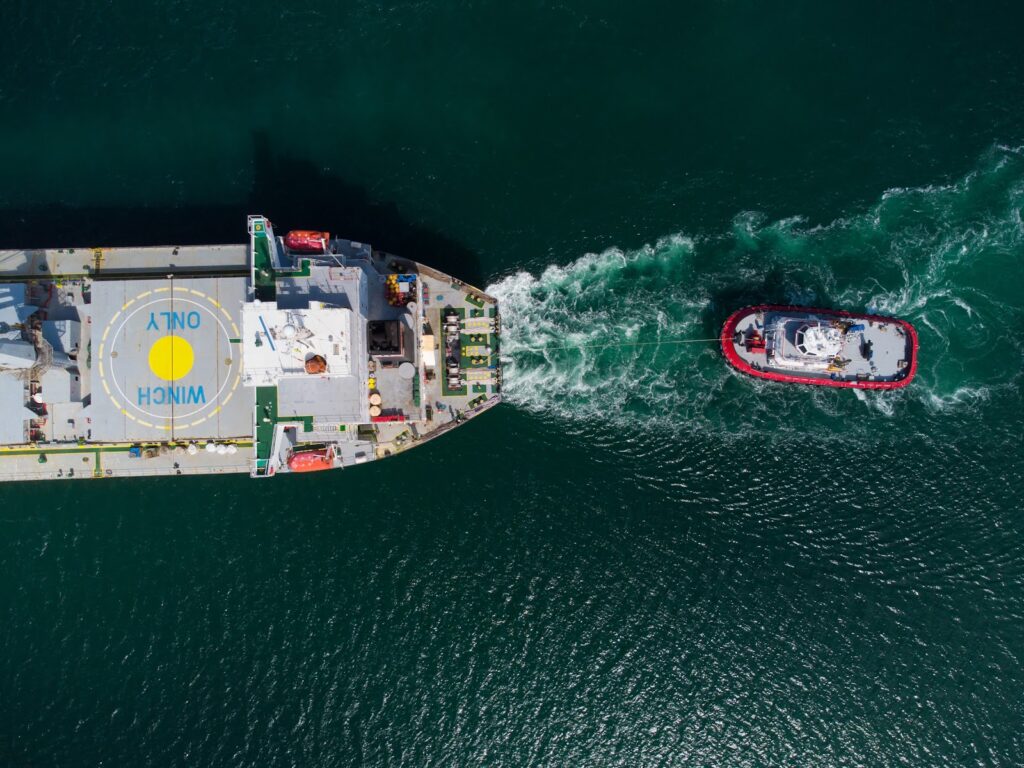 Aerial view of a tugboat towing a large ship with a helipad marked “WINCH ONLY” on its deck, both moving through deep green water, leaving visible white wakes behind them.