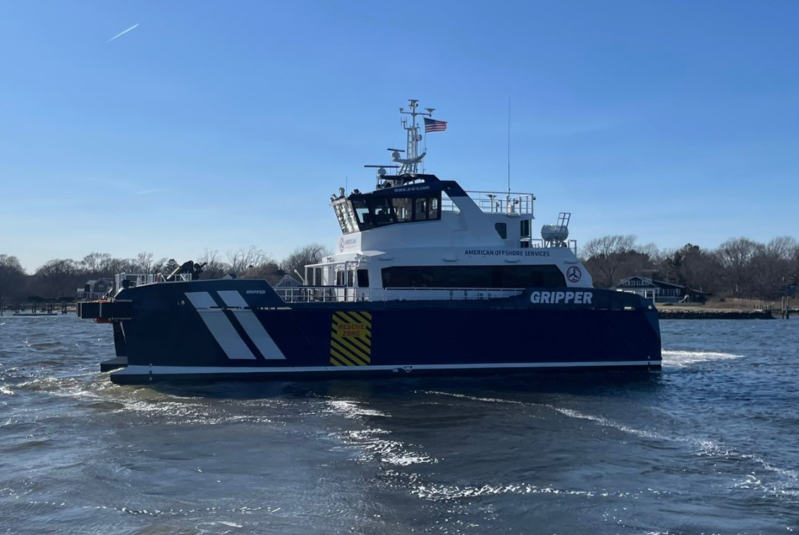 A blue and white vessel named "GRIPPER" with American Offshore Services branding moves across a body of water on a sunny day, with trees and houses visible in the background.