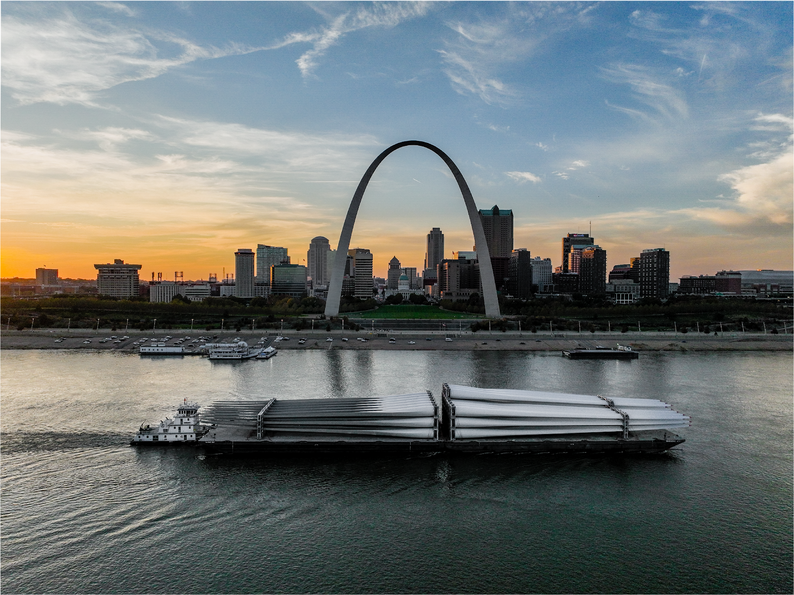 A barge carrying large wind turbine blades passes on the Mississippi River in front of the Gateway Arch and downtown St. Louis at sunset, with city buildings silhouetted against the sky.