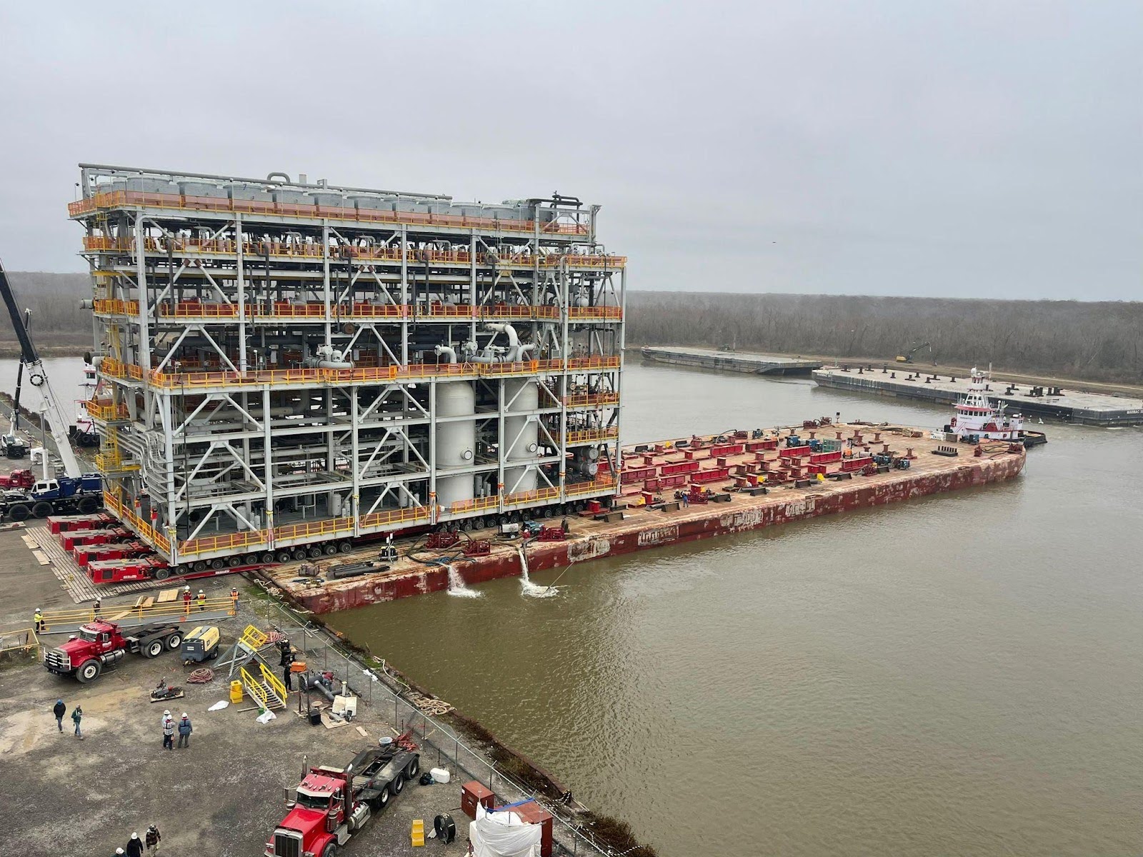 A large industrial module is being loaded onto a barge at a riverside dock. Several trucks, workers, and equipment are visible nearby; a tugboat waits in the water beside the barge under an overcast sky.