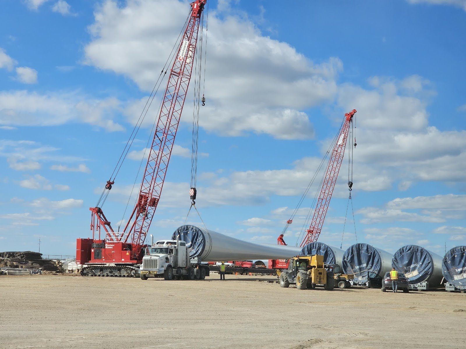 Two large red cranes lift and move sections of wind turbine towers at a construction site, with several turbine sections lined up on the ground under a blue sky with scattered clouds.