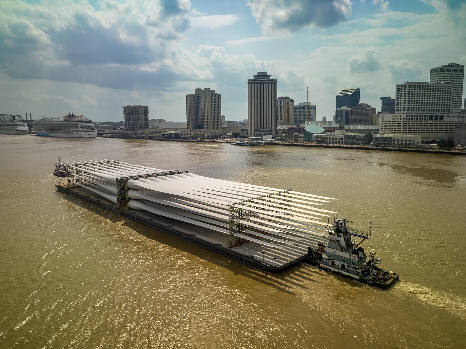 A barge loaded with large wind turbine blades is pushed by a tugboat on a wide brown river, with a city skyline and tall buildings visible in the background under a partly cloudy sky (RiverLens).