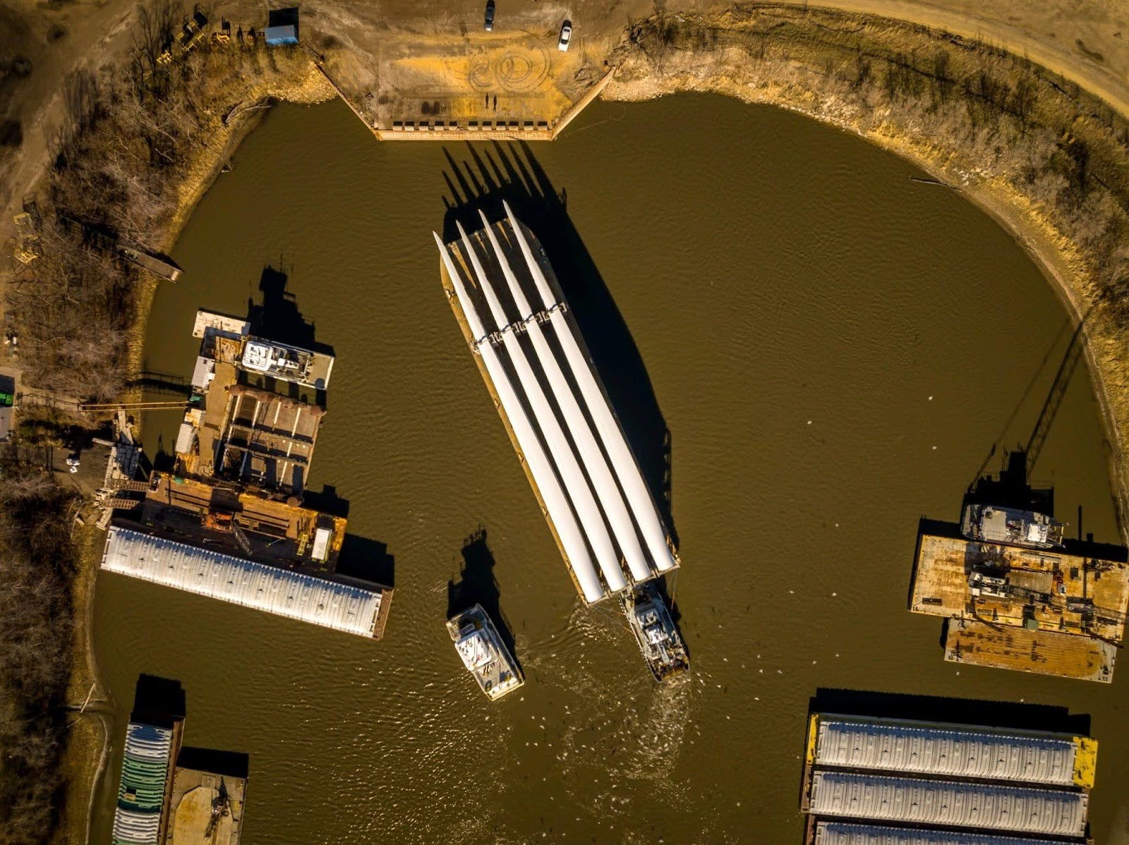 Aerial view of a barge carrying three large wind turbine blades on a brown river, surrounded by docks and industrial structures. Tugboats guide the barge while the shadows of the blades extend across the water.