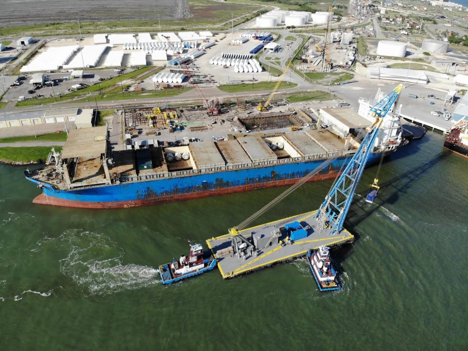 Aerial view of a large blue cargo ship docked at an industrial port, with a crane on a barge lifting heavy equipment from the water, and storage tanks and buildings visible in the background.