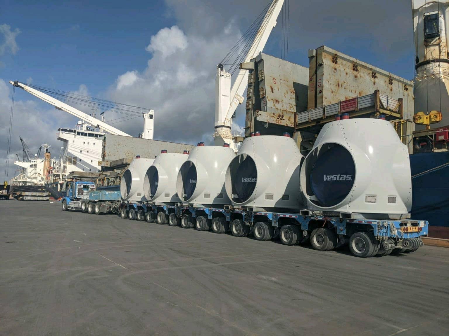 A long flatbed truck carries five large white Vestas wind turbine nacelles, lined up at a busy industrial port with cranes, ships, and containers in the background under a partly cloudy sky.