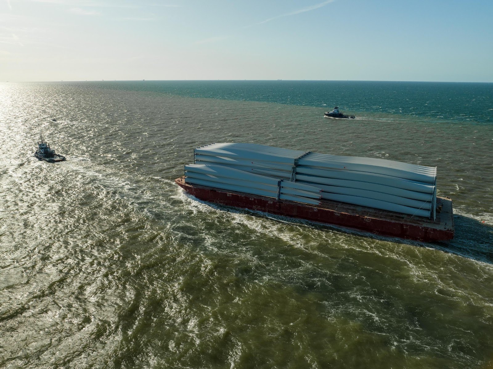 A barge carries stacked wind turbine blades across a wide expanse of sea, accompanied by two tugboats under a clear blue sky.