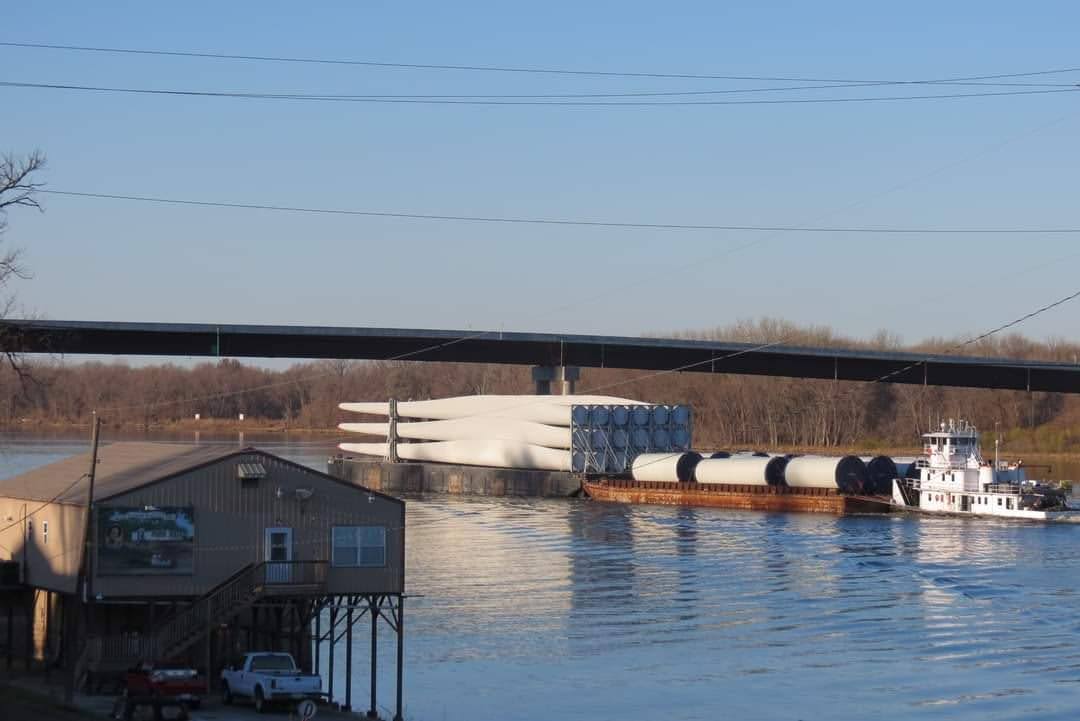 A barge transports several large wind turbine blades and cylindrical components on a river, passing a riverside building and vehicles, with a bridge and trees in the background under a clear sky.