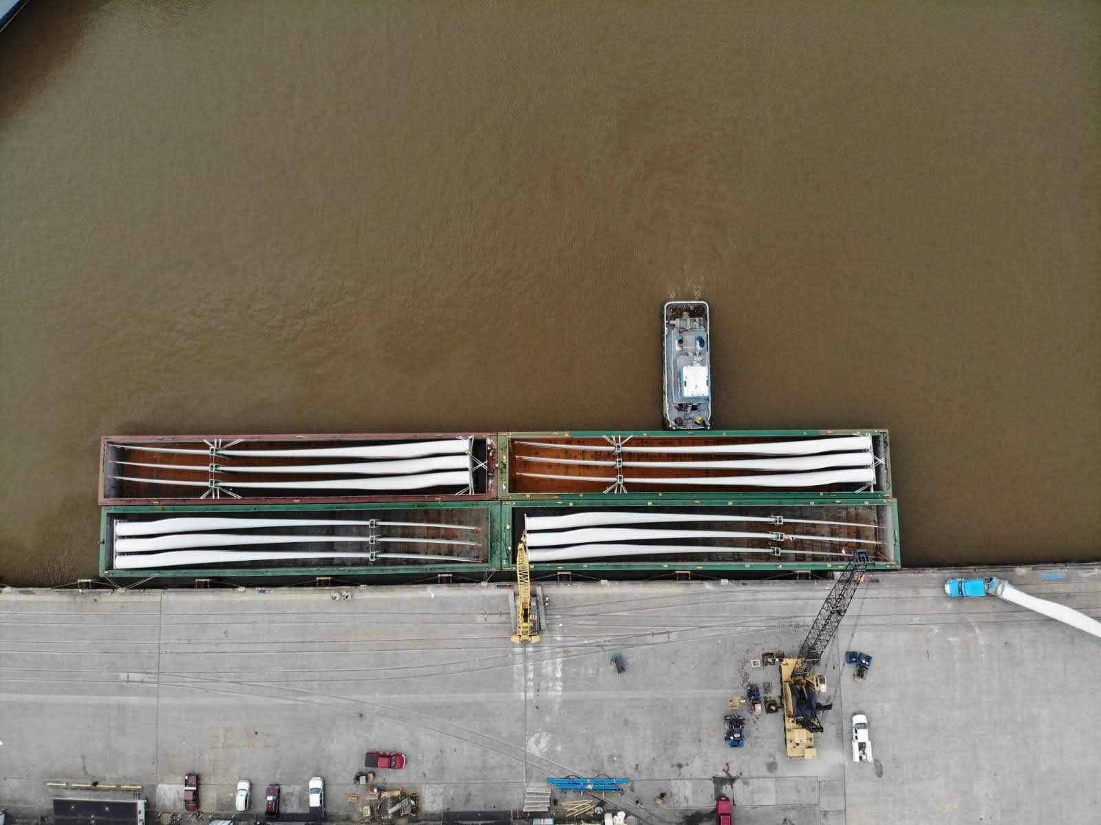 Aerial view of a dock where large wind turbine blades are loaded onto a barge by cranes. Vehicles and machinery are positioned along the edge of the concrete dock next to the brown river.
