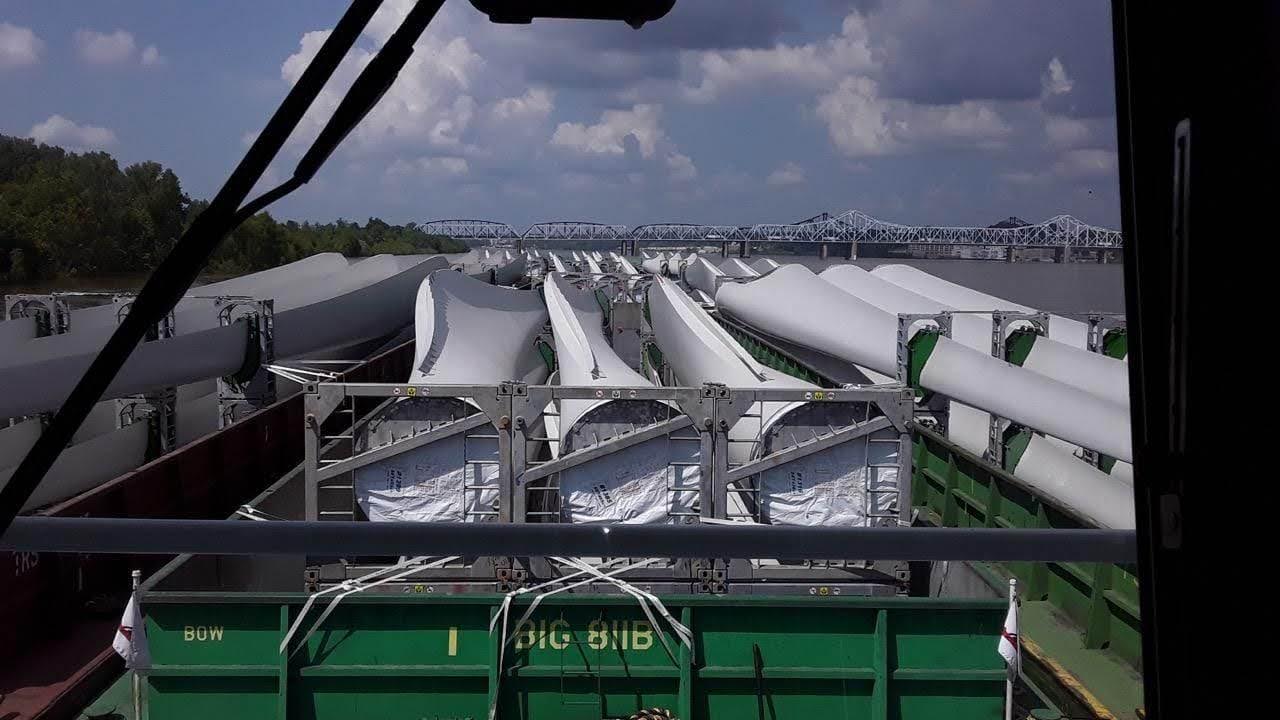A view from a boat's cabin shows large wind turbine blades stacked on barges floating on a river, with a bridge and trees visible in the background under a partly cloudy sky.