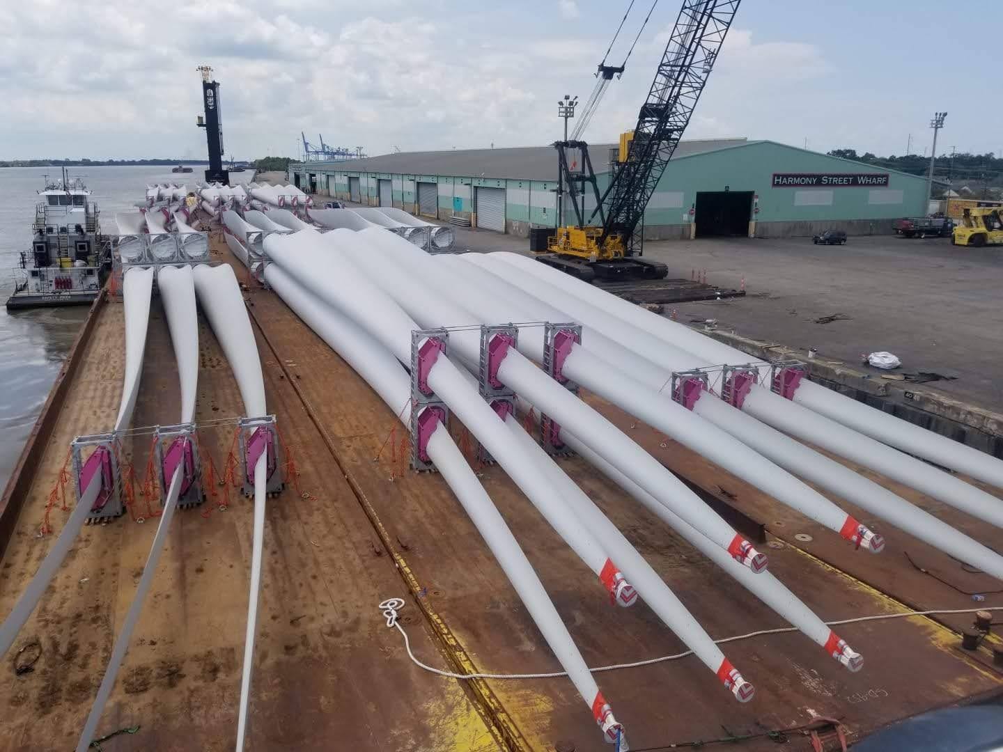 A large barge docked at a wharf is loaded with multiple long white wind turbine blades, neatly lined up in rows. Cranes and industrial buildings are visible in the background.