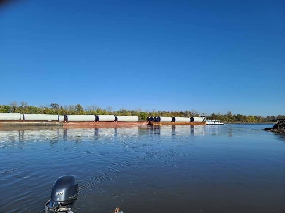 A long barge carrying large cylindrical objects floats on a calm river under a clear blue sky, with trees lining the distant riverbank and a boat motor visible in the foreground.