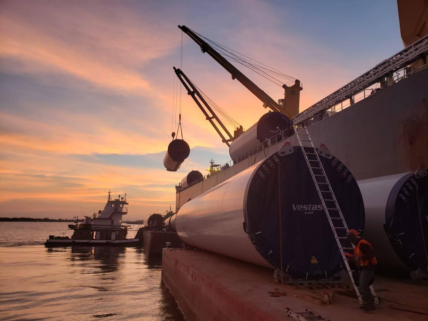 A crane on a docked ship lifts a large cylindrical object at sunset. Wind turbine blades labeled "Vestas" are stacked nearby, with a worker in a safety vest and helmet standing on a ladder. A tugboat is on the water.