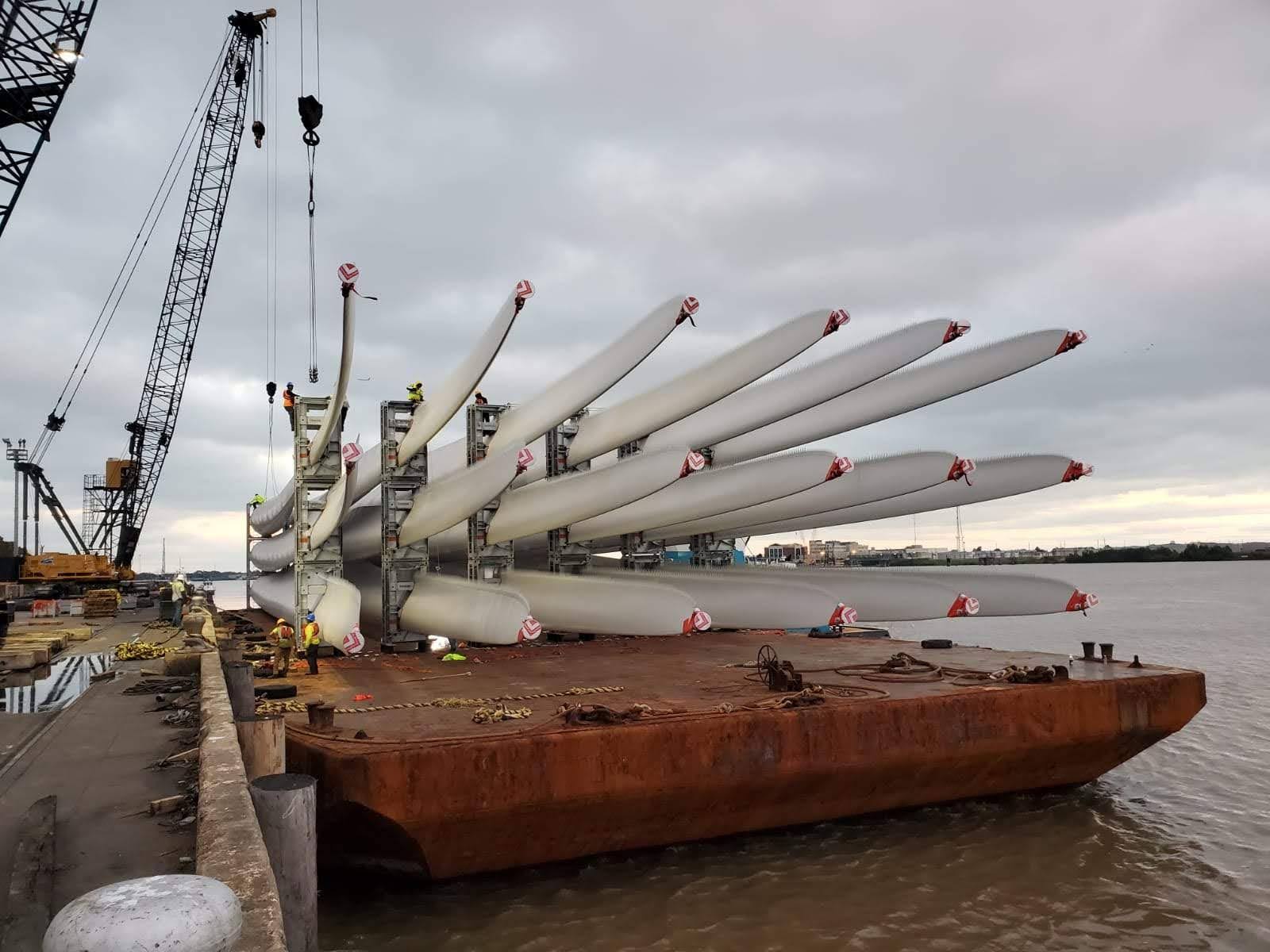Large wind turbine blades are stacked and secured on a flat barge at a dock, with cranes and workers nearby, under a cloudy sky by the water.