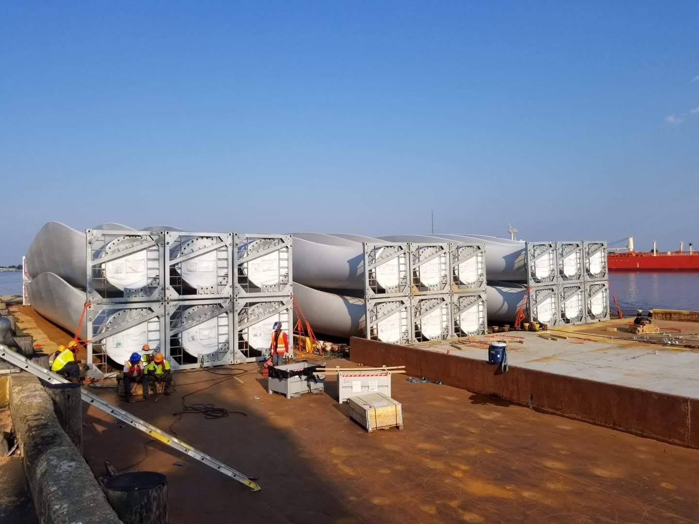 Large wind turbine blades are stacked horizontally in metal frames on a dockside, with workers in safety gear nearby and a ship in the background under a clear blue sky.