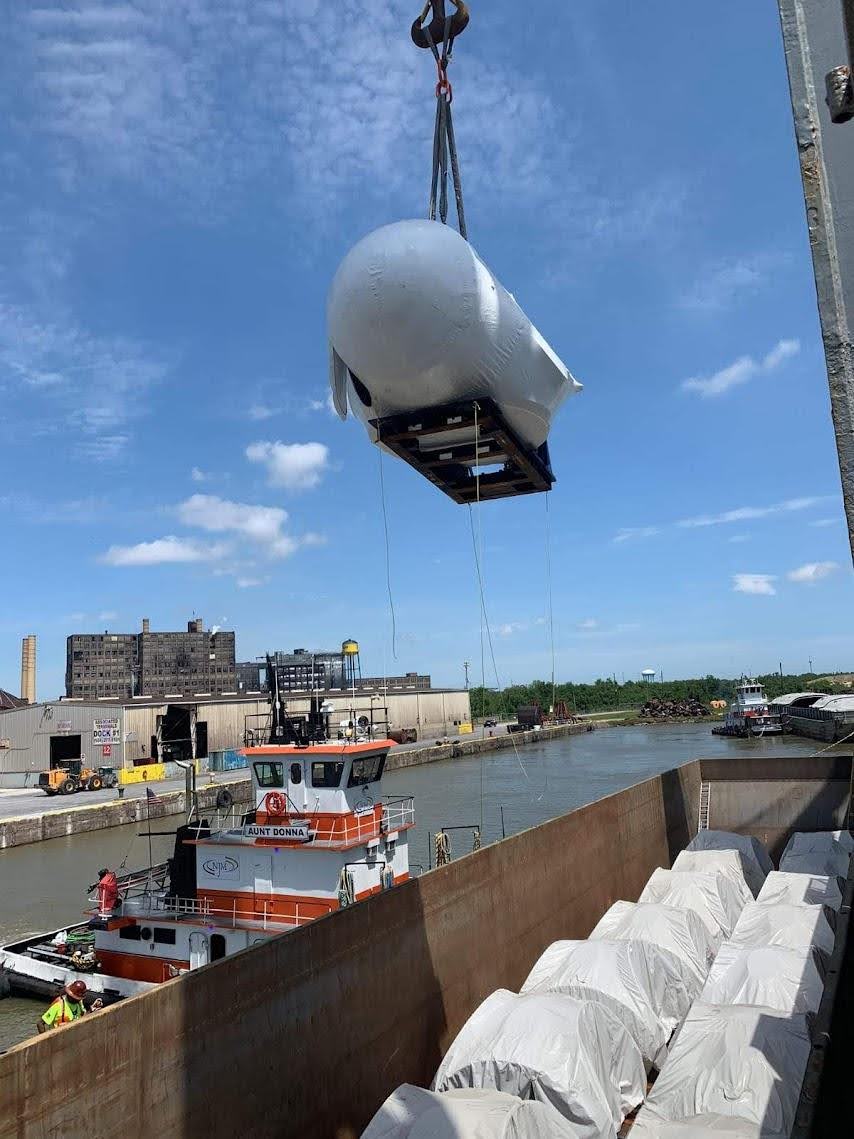 A large industrial object is being lifted by a crane over a dockside canal, with a tugboat and covered cargo in the foreground and industrial buildings in the background under a blue sky.