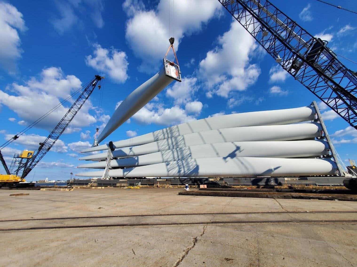 Large wind turbine blades are stacked horizontally on the ground, while a crane lifts one blade. Another crane is nearby under a blue sky with scattered clouds at an industrial site.