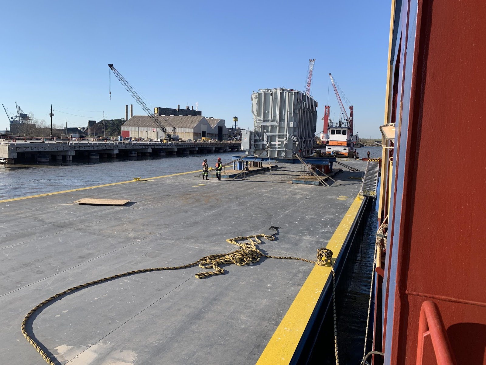 Workers stand on a large barge with a heavy industrial object secured on deck; cranes and buildings are visible along the waterfront under a clear blue sky.