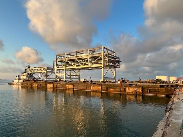 A large, metal industrial structure sits on a barge in calm water under a partly cloudy sky, with a tugboat nearby and additional industrial equipment visible in the background.