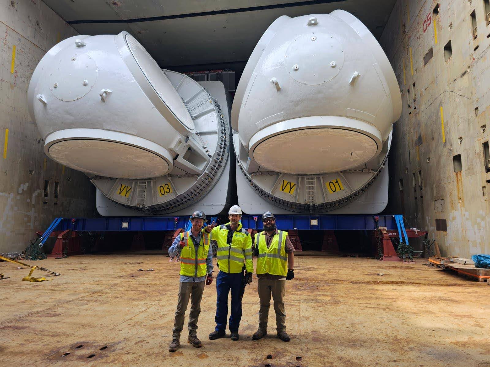 Three workers in safety vests and helmets stand in front of two massive white industrial structures inside a large, open facility, posing for a photo and giving a thumbs-up gesture.
