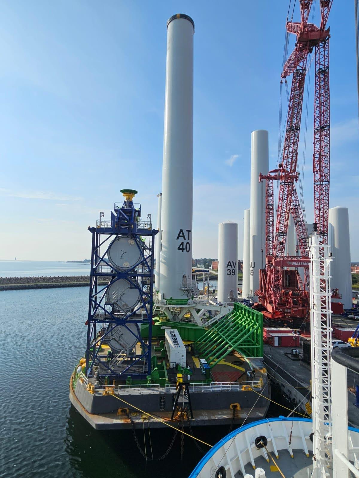 A large offshore wind turbine tower section stands vertically on a dock next to a red crane and other tower sections, with a specialized transport barge floating in the harbor. The sky is clear and calm.