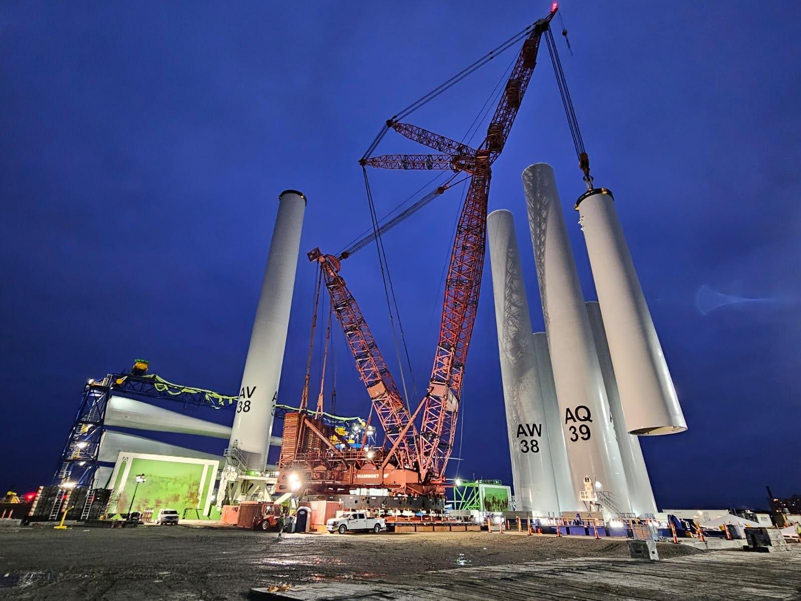 A large crane lifts a massive cylindrical wind turbine tower section at an industrial construction site at night, surrounded by other tower sections and construction equipment under a dark blue sky.