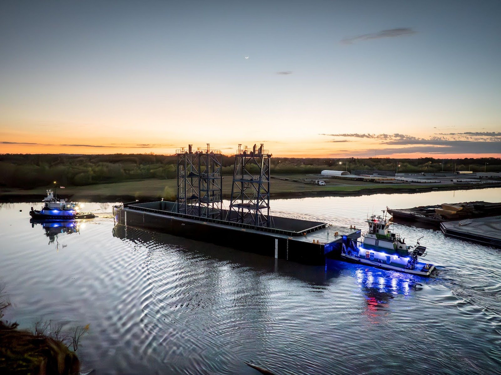 Two tugboats guide a large industrial barge with tall metal structures down a calm river at sunset, with blue lights reflecting on the water and a scenic landscape in the background.