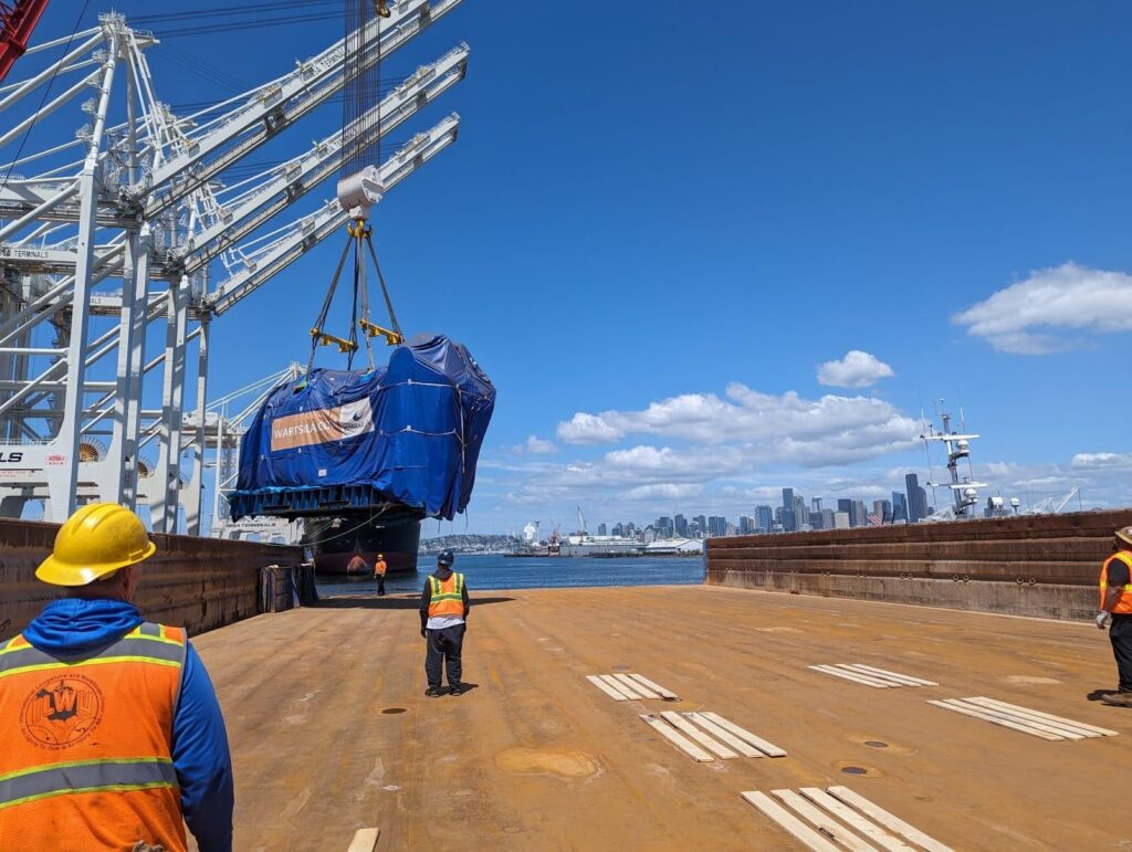 Workers in safety gear guide a large, tarp-covered object being lifted by a crane onto a cargo ship at a port, with a city skyline and blue sky with clouds in the background.