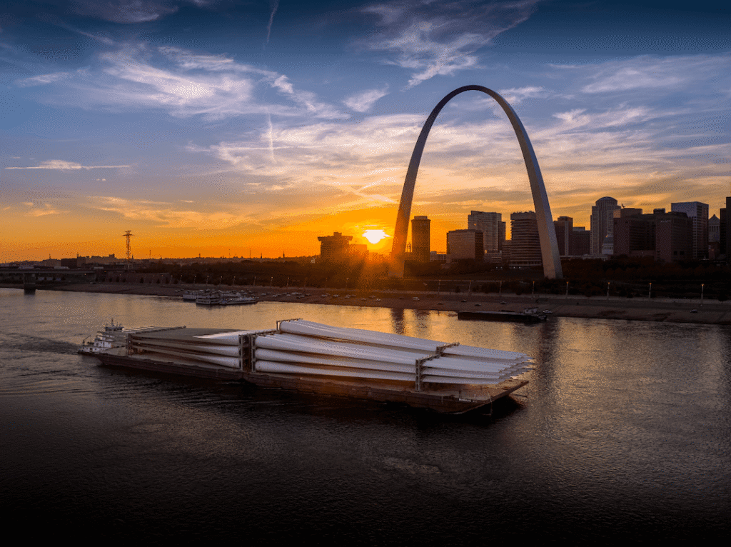 A barge carrying large cylindrical objects travels along the river at sunset, showcasing inland waterway transport with Riverlens, with the St. Louis Gateway Arch and city skyline silhouetted in the background.