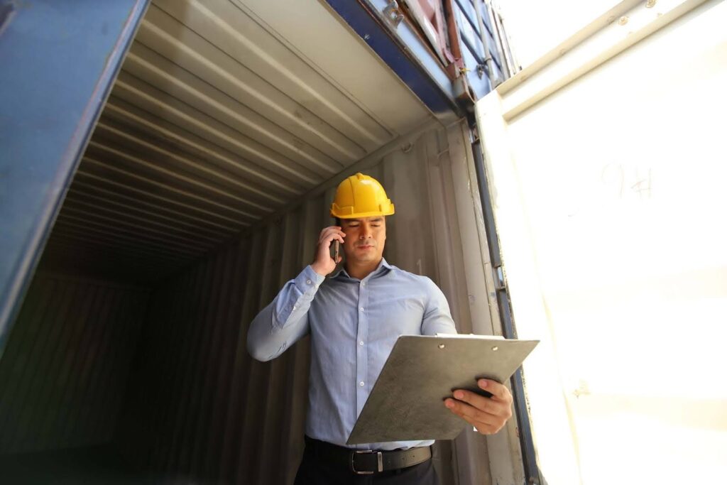 A person wearing a yellow hard hat stands inside a shipping container, talking on a phone and holding a clipboard, appearing to check project logistics documents during an inspection.