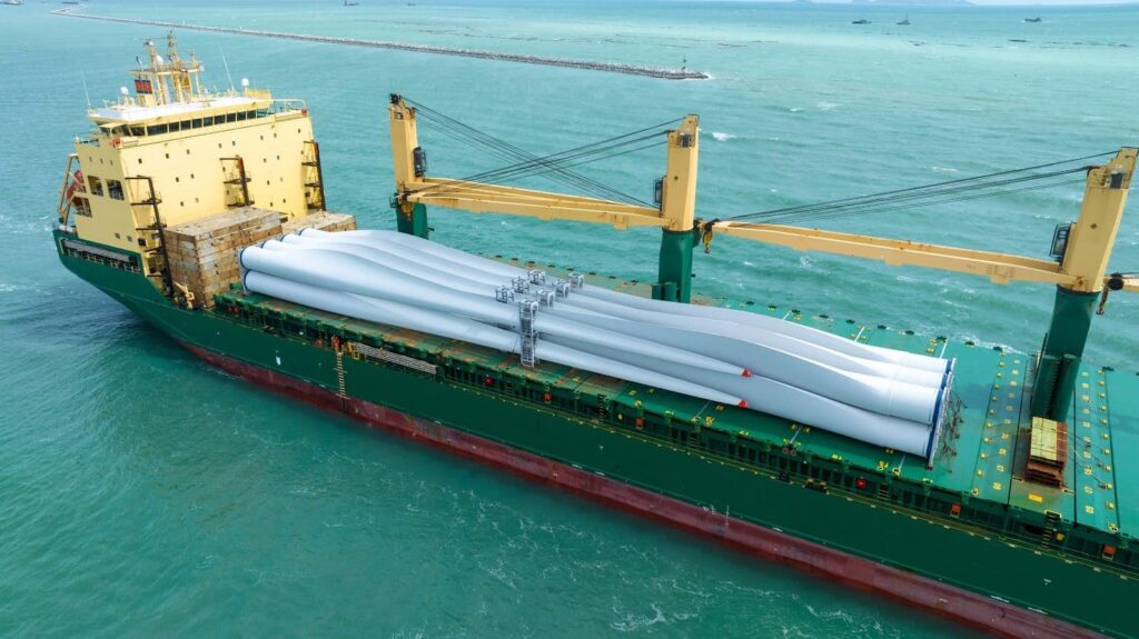 A large green cargo ship, managed by project logistics experts, transports several long, white wind turbine blades across turquoise waters, with the coastline and other vessels visible in the distance.