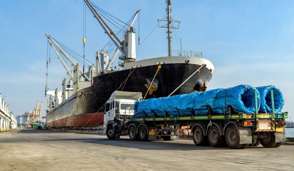 A large cargo ship is docked at a port, with cranes overhead. In the foreground, a truck—managed by project logistics experts—carries large cylindrical items covered in blue tarps on a flatbed trailer under the clear blue sky.