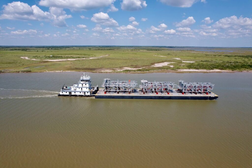 A large barge carrying industrial equipment is being pushed by a tugboat on a wide river, overseen by project logistics experts, with grassy plains and a partly cloudy sky in the background.