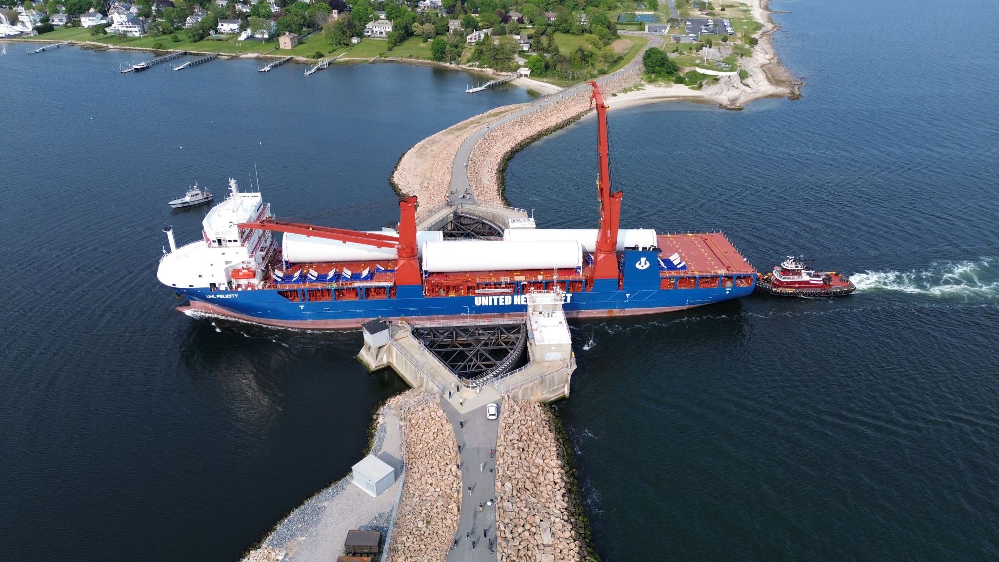 A large cargo ship, assisted by a tugboat, passes through a narrow canal with stone breakwaters and a raised drawbridge. Houses and greenery line the distant shoreline under a clear sky.