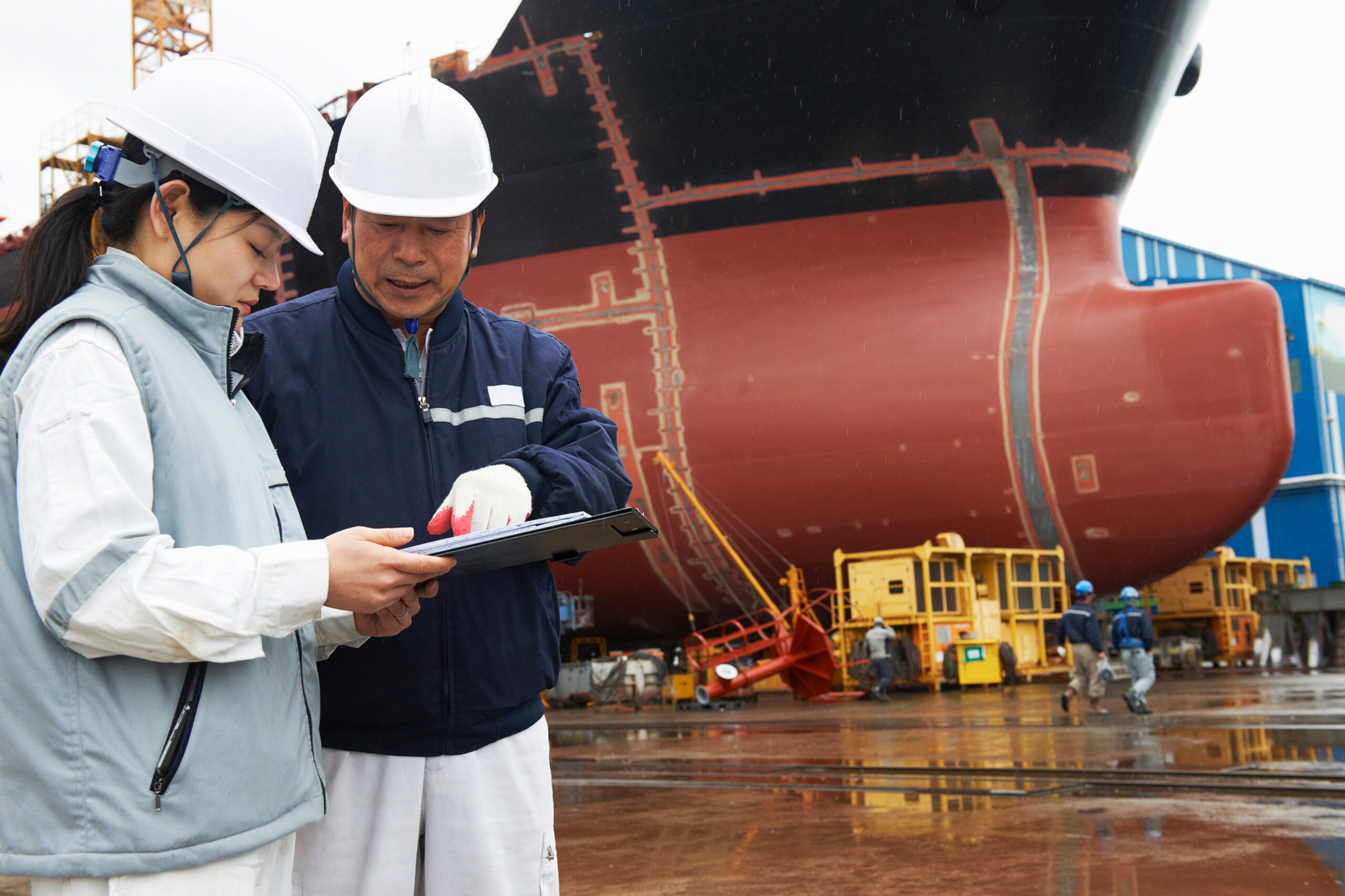Two people wearing white hard hats and work jackets review a clipboard in a shipyard, standing in front of a large ship under construction. The ground is wet and equipment is visible in the background.