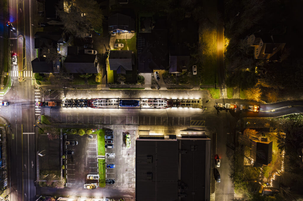 Aerial night view of a brightly lit street with parked cars, buildings, and houses. Streetlights illuminate the road and parking lot, highlighting areas popular with heavy haul trucking in this bustling residential neighborhood.