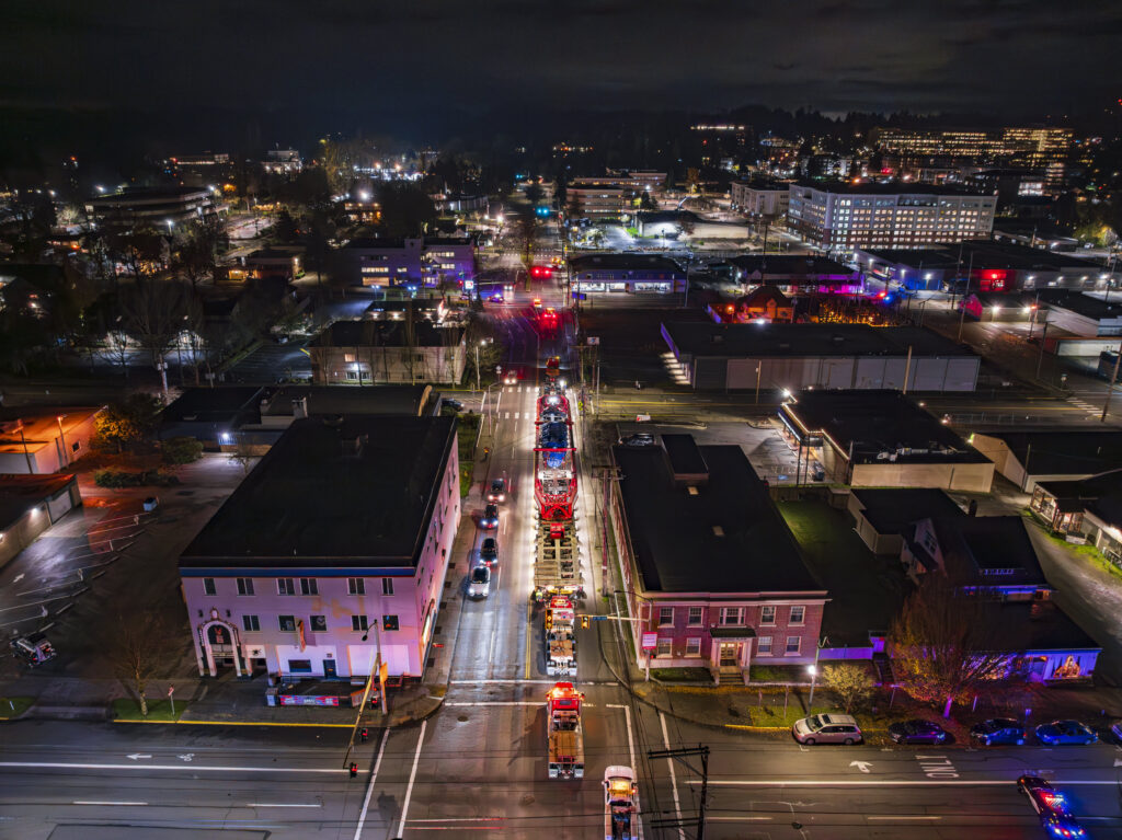 A nighttime aerial view of a city street lined with fire trucks displaying flashing lights, surrounded by buildings and illuminated streets, highlights the cityscape and hints at the scale required for heavy haul logistics in urban environments.