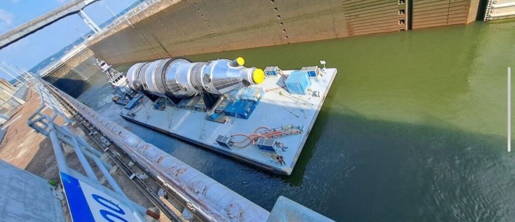 A large industrial cylinder, transported by SPMT and secured on a barge, is floating through a lock system surrounded by high concrete walls and bridges overhead, with clear skies in the background.