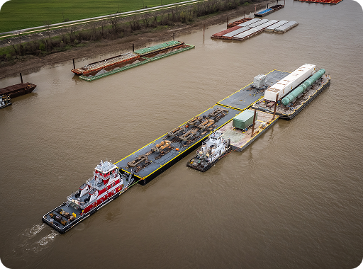 A red and white tugboat pushes several barges carrying industrial cargo along a wide brown river, with more barges and a green field visible in the background.