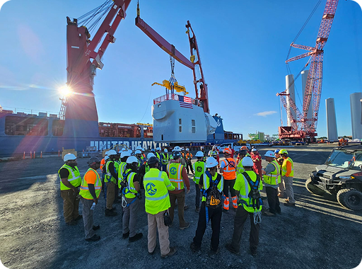 A group of construction workers in safety gear gather near cranes lifting a large industrial component at a port, with wind turbine parts and machinery in the background under a clear blue sky.