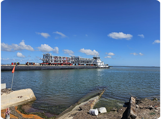 A large industrial barge loaded with equipment travels along a wide, calm waterway under a clear blue sky with scattered clouds. In the foreground, there’s debris and concrete on the shore.