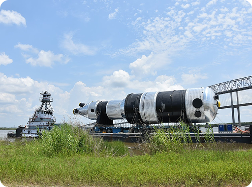 A large black and silver rocket section is transported on a barge along a river, with a tugboat beside it and a bridge visible in the background under a partly cloudy sky.