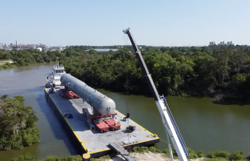 A large cylindrical tank, transported on a barge with a tugboat, awaits transfer onto a self propelled modular transporter (SPMT) by a crane extending over the water, with dense trees and industrial buildings in the background.