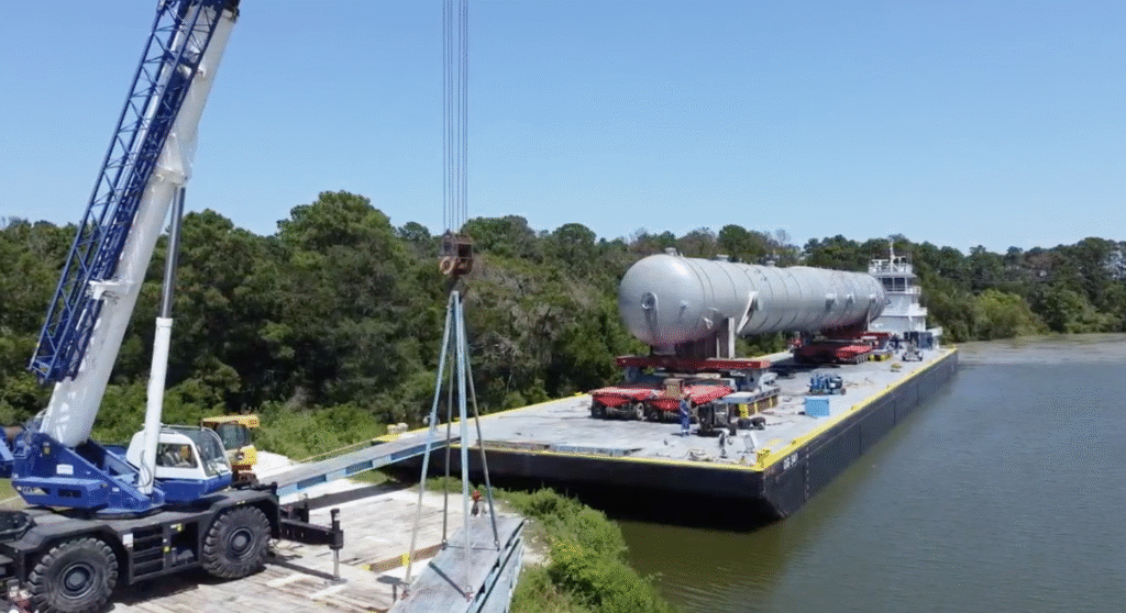 A large cylindrical industrial tank is being transported on a barge along a waterway, with heavy loads secured on board and vehicles including an SPMT visible, while a crane operates nearby under a clear blue sky.