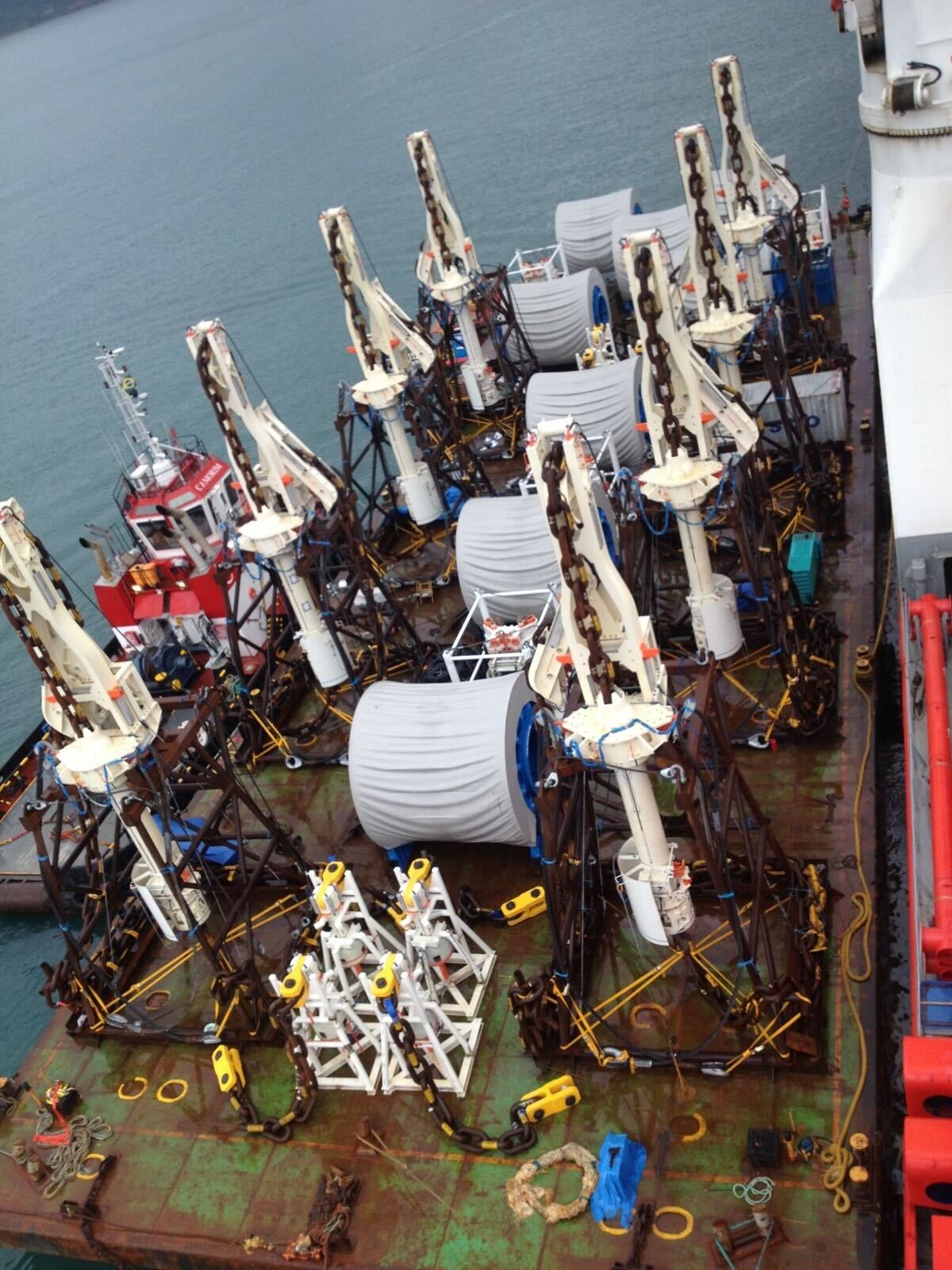 A top-down view of a ship deck loaded with large industrial equipment, including multiple white cranes and large cable reels, docked beside a red support vessel on the water (offshore marine logistics).
