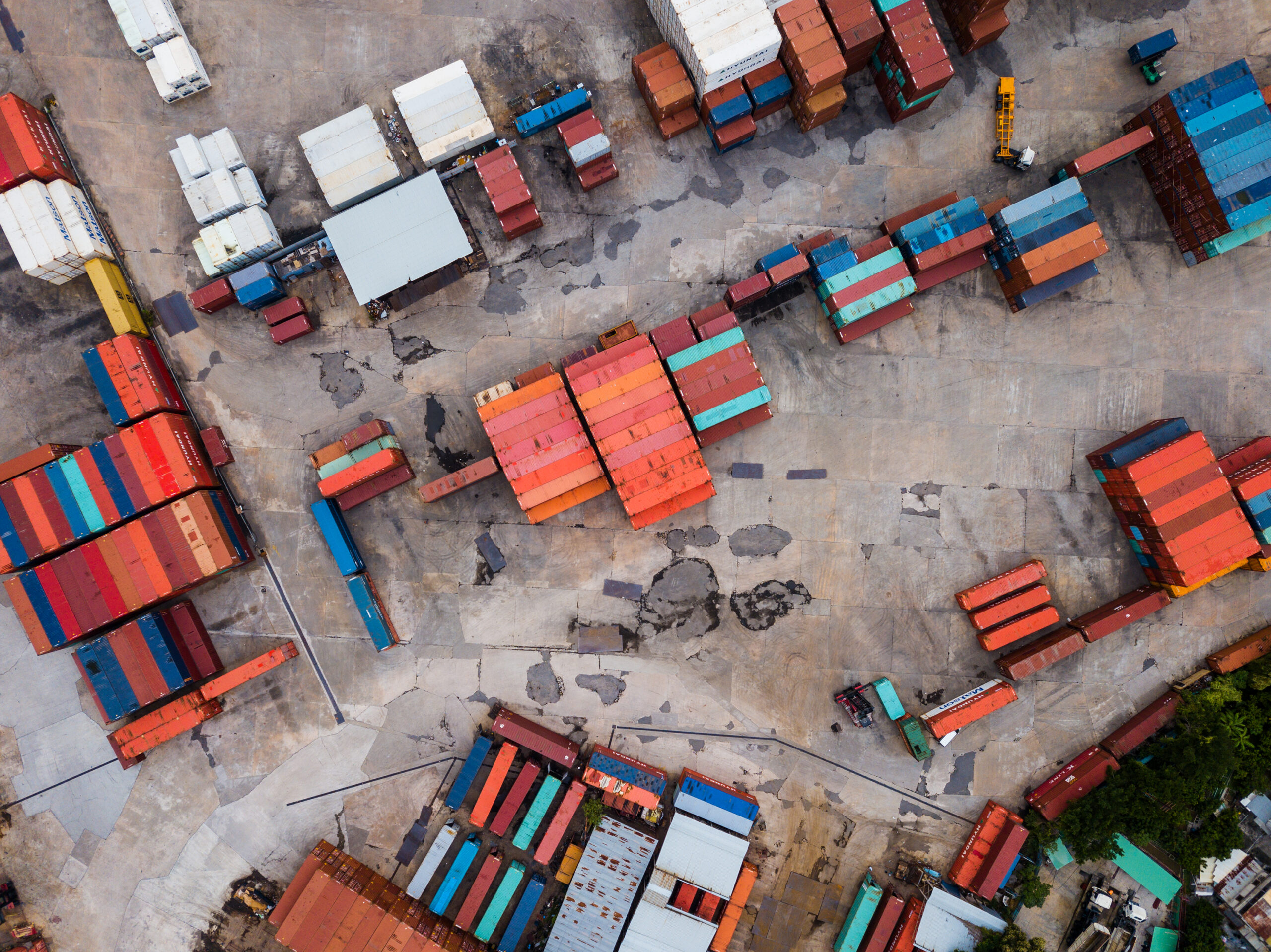 Aerial view of a shipping yard with rows of colorful cargo containers organized on a concrete surface, interspersed with trucks and small buildings.