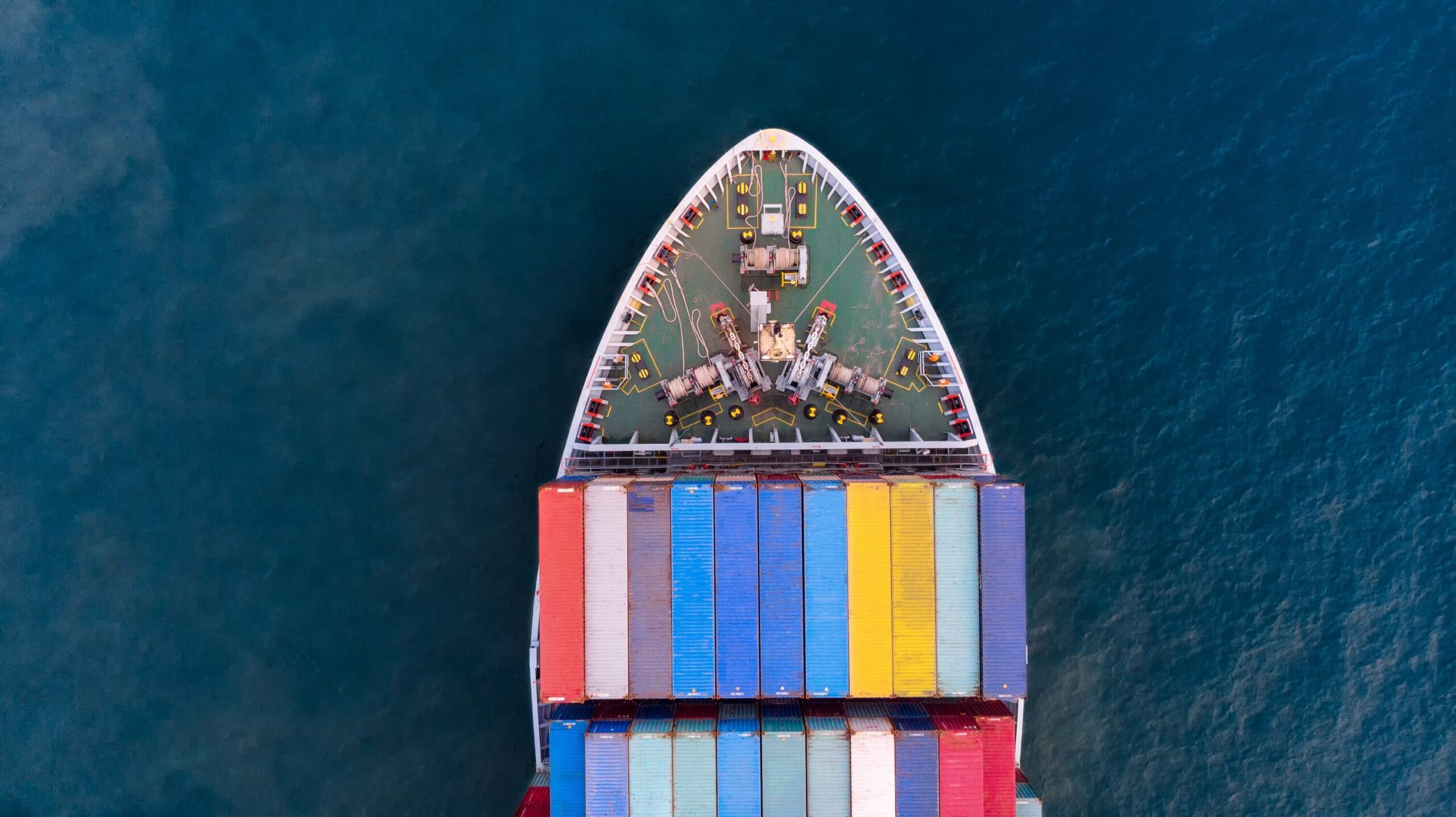 Aerial view of the bow of a cargo ship carrying colorful stacked shipping containers, sailing on deep blue water after vessel clearance.